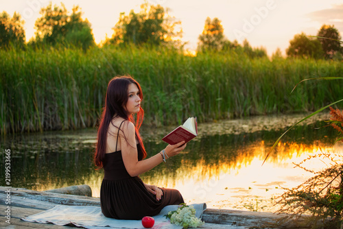 Young woman reading a book on a wooden pier by the river during a beautiful golden hour sunset