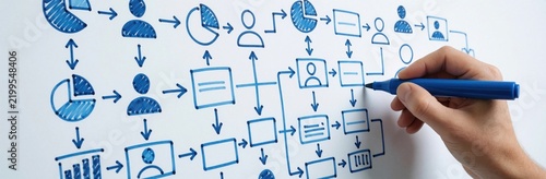 Hand draws complex flowchart on whiteboard with blue marker. Visualizes business process stpic, data flow, decision trees. Charts depict project phases, user journeys, team workflows. Strategic