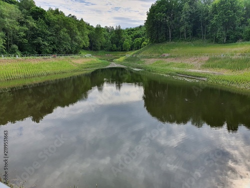 A serene landscape view of a calm water reservoir or pond nestled within a lush green environment. The water surface mirrors the partly cloudy sky and surrounding dense forest, creating a