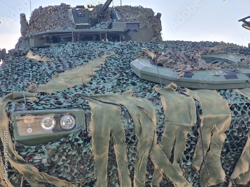 Konstfotografi Close-up view of a modern military armored combat vehicle, likely a main battle tank or infantry fighting vehicle, extensively covered with tactical camouflage netting and fabric strips
