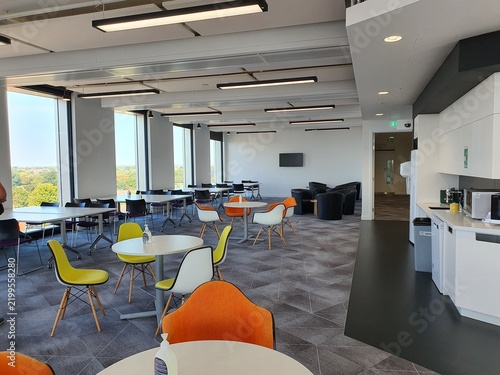 A modern, spacious office break room or cafeteria featuring floor-to-ceiling windows with views of green trees outside. The interior includes multiple white round and rectangular tables surrounded