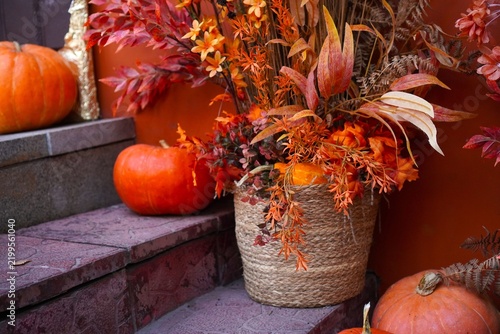 Orange pumpkins and autumn flowers arrangement in woven basket decorating home porch steps for fall harvest festival and Halloween. Seasonal porch decor with warm orange color palette.