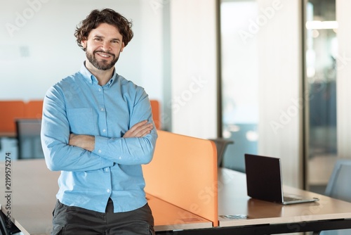 Konstfotografi Smiling Professional White Male Businessman With Crossed Arms In Office Setting, Enjoying Work Environment, Standing Next To Desk With Laptop