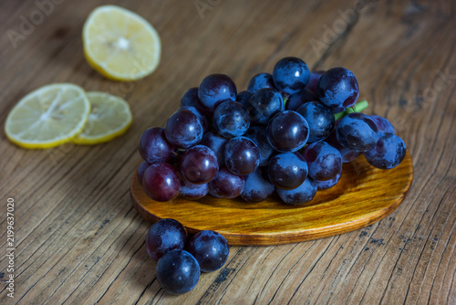 A cluster of dark purple grapes rests on a small wooden plate, with lemon slices placed nearby on a wooden table surface.