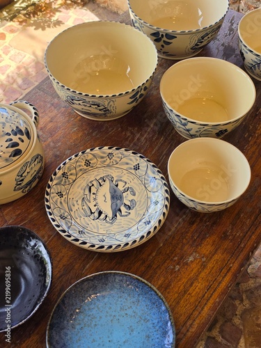 Handmade cream ceramic bowls and plates with hand-painted blue crab and floral motifs sit on a rustic wooden table, including a lidded tureen and smaller serving pieces. The collection has a warm, art