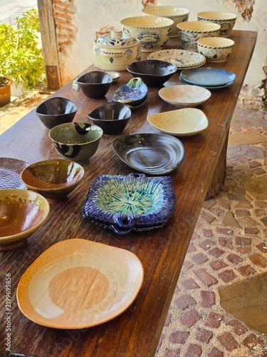 Handmade cream ceramic bowls and plates with hand-painted blue crab and floral motifs sit on a rustic wooden table, including a lidded tureen and smaller serving pieces. The collection has a warm, art