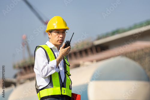 Bild auf Leinwand Asian construction engineer in hard hat and safety vest talking walkie talkie on