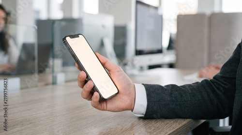 Businessman holding blank screen smartphone mockup in modern office workspace, professional using mobile device for work presentation or communication in corporate environment.