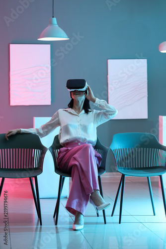 Young woman with VR glasses sitting in modern art gallery
