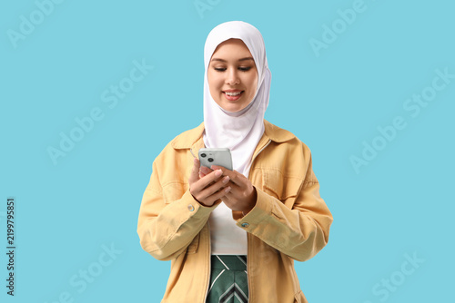 Beautiful young Muslim woman in hijab with mobile phone on blue background