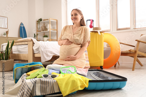 Young pregnant woman with clothes and travelling accessories packing suitcase...