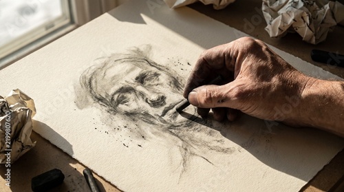 Artist hand sketching a detailed human portrait using charcoal on textured paper.