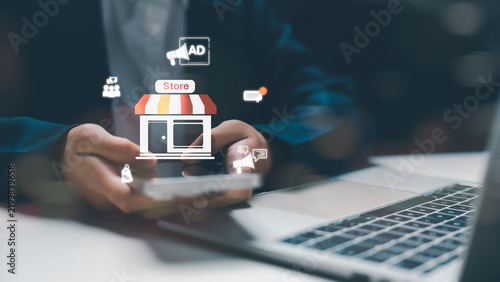 Male businessman using smart phone with digital store icon and laptop for online shopping retail and social media advertising campaign concept in modern corporate office desktop workspace.