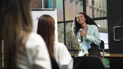 Smiling businesswoman giving presentation with microphone to colleagues in modern office, sharing ideas during corporate training, team meeting and professional workplace communication.