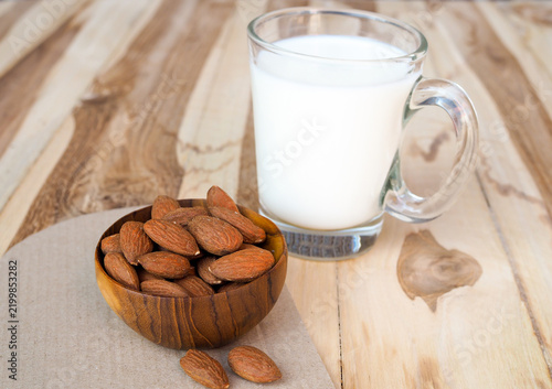 Glass of fresh almond milk with raw almonds in wooden bowl on wooden background.