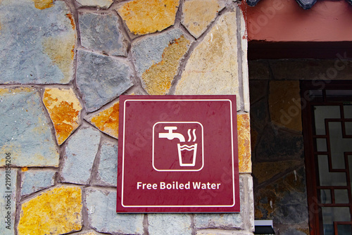 Brown sign offering free boiled drinking water, mounted on rustic stone wall. Sign features water glass icon and clear text, representing public access to clean drinking water community hospitality.