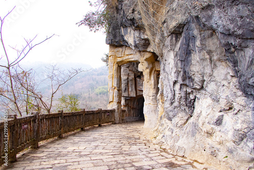 Scenic stone walkway along dramatic limestone cliff leading through natural cave entrance with wooden railing mountain landscape in background. Hiking trail surrounded by rugged rock formations nature