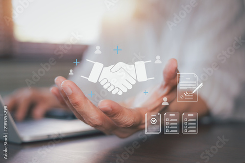 Businessman hand presenting virtual handshake hologram with contract documents. Concept of digital partnership, online agreement, business deal, trust, and corporate teamwork collaboration.