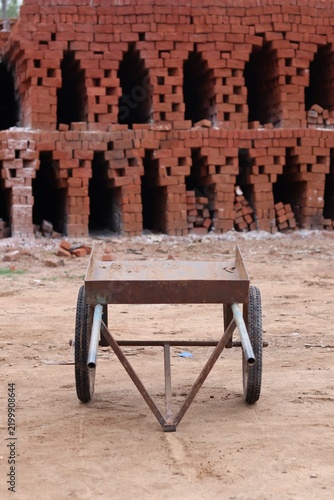 Traditional Metal Wheelbarrow at a Rural Brick Kiln with Stacked Red Clay Bricks in India.