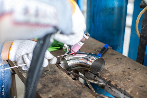 Selective focus to hands of worker positioning an argon TIG welding torch and filler rod on a polished stainless steel pipe elbow in a workshop.