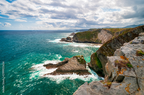 Breathtaking rugged coastline with turquoise ocean waves crashing against limestone cliffs and a rocky sea stack beneath a dramatic cloudy sky, evoking wild, untamed coastal beauty and adventure.