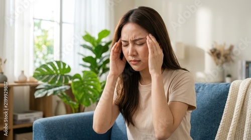 Young Asian woman suffering from severe headache or migraine while sitting on sofa at home, feeling stressed and unwell.