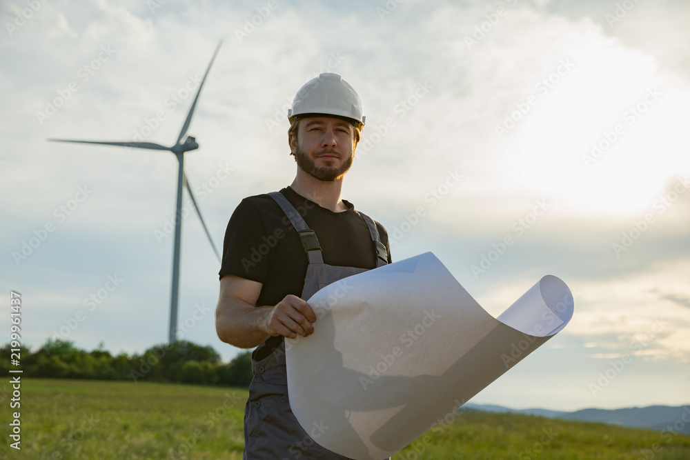 Obraz premium A Caucasian man in work clothes and hard hat stands in a grassy field with a wind turbine behind him, holding rolled-up plans