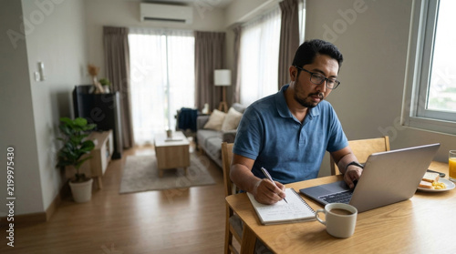 Southeast asian man working remotely on a laptop and taking notes at a dining table in an apartment