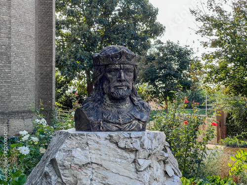 Bronze bust of crowned Saint Stephen with beard and long hair on a rough stone pedestal, surrounded by red and white roses and green trees near a brick church in Čantavir, Serbia, September 2026.