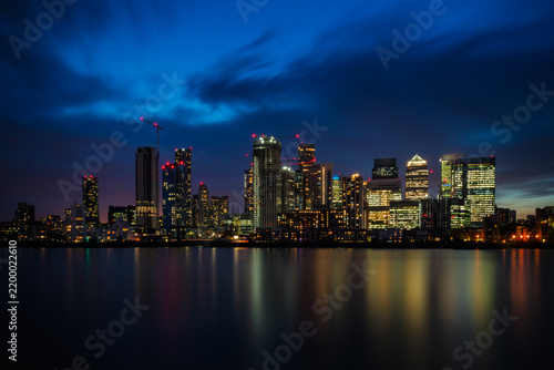 An illuminated skyline of Canary Wharf with colourful reflections shimmering across the River Thames in London, England