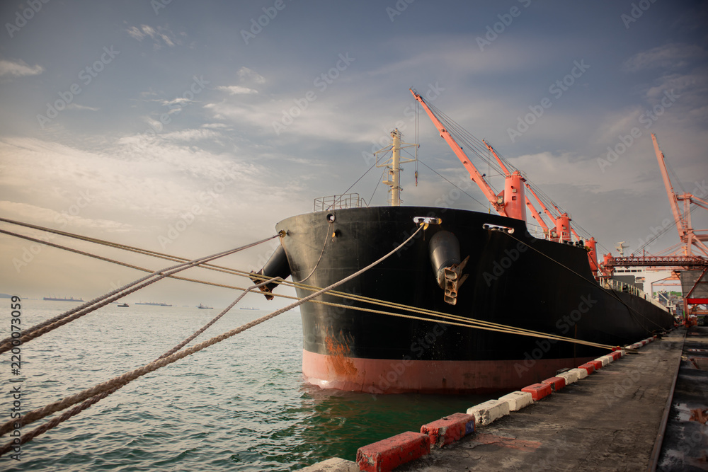 Naklejka premium Cargo ship moored at industrial harbor with orange crane, heavy rope, calm seawater, and dramatic cloudy sky creating maritime transport scene
