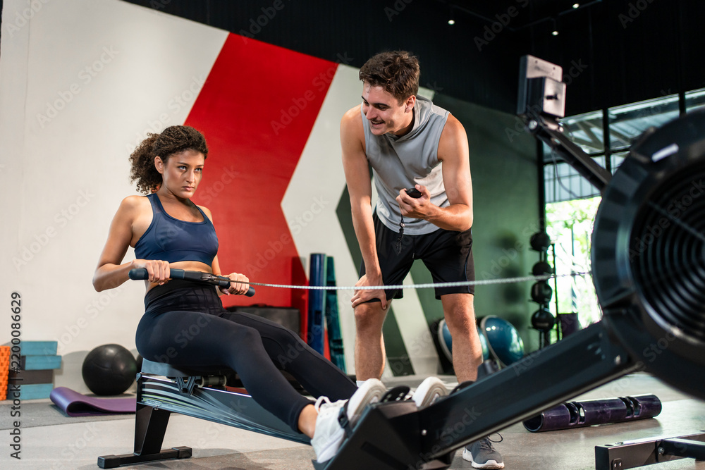 Obraz premium Young athletic woman using rowing machine with trainer in fitness gym. 