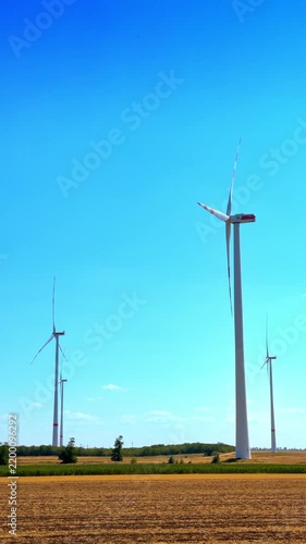 Tall wind turbines in rural field against clear sky. Several white wind turbines with red stripes on blades stand in a dry harvested farm field during daytime.