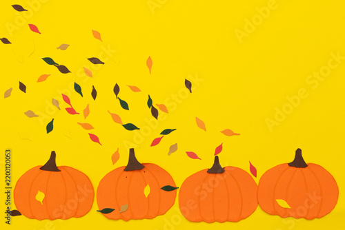 Paper pumpkins with small leaves in warm tones falling onto them against a yellow background, concept of the arrival of fall and seasonal transition