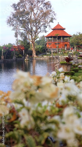 Ornate pavilion stands by garden pond amid tropical greenery. Red roof building, tall tree and decorative statues surround tranquil water, with blurred flowers adding soft foreground depth.