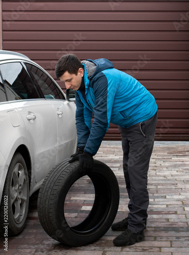 A man is rolling a black car tire near a white vehicle for seasonal wheel replacement