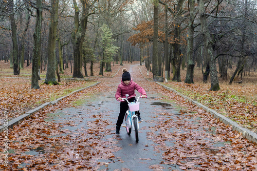 Happy little girl riding a bicycle on a wet park path covered with fallen leaves during a crisp autumn day. Autumn walk in the park