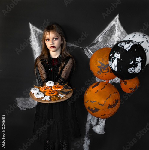 A little girl dressed as a vampire is holding a tray of Halloween cookies. Vampire girl on a black background. Halloween concept
