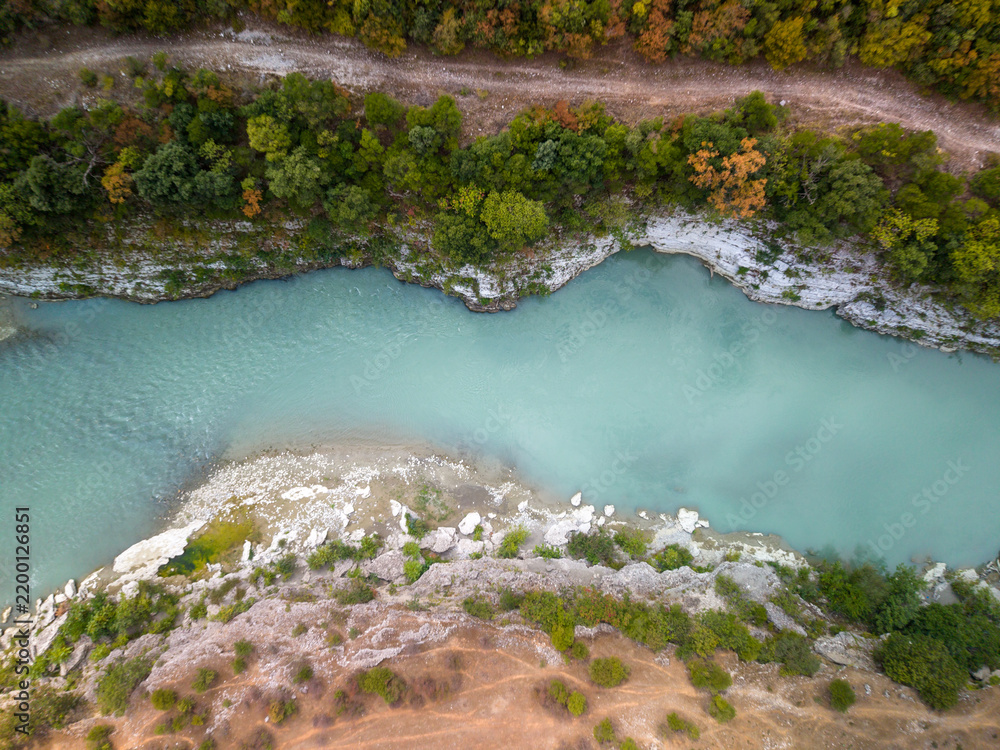 Fototapeta premium Vjosa River in Albania flows beneath top down aerial perspective, bordered by chalky cliffs and green woodland. Calm turquoise channel gives this remote river landscape peaceful, scenic mood.