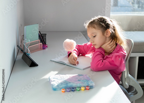 A focused little girl sits at the desk and carefully writing in her notebook while studying at home during an online class on a tablet. Concept of school, homework and the beginning of the school year