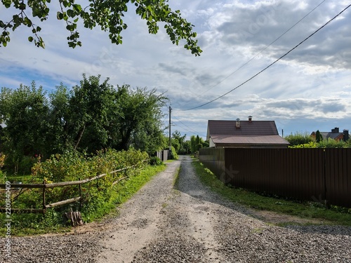 фотографія On one side of the street, strewn with rubble, bushes and fruit trees grow behind wooden fences, and on the other, living houses stand behind metal fences
