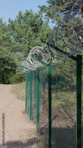 High razor wire coils top a green chainlink fence stretching along a sandy path by pine trees under daylight. Concept of perimeter security, border defense, and area containment.