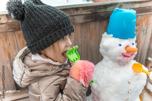 Bild auf Leinwand Cute curious little girl in black knitted beanie hat playfully tasting snow from