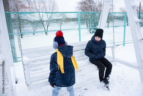 Two children bundled in winter wear sit on a snowy swing set, gazing out at a frosty landscape.