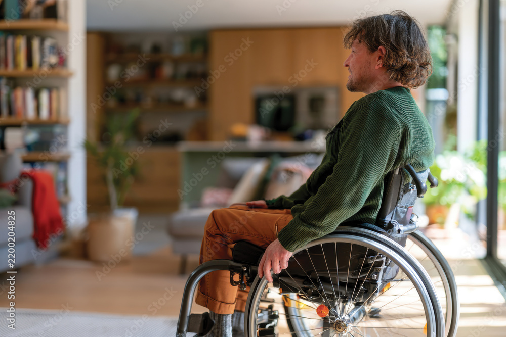 Obraz premium A thoughtful man sits in a wheelchair at home, reflecting in a bright living space, portraying comfort and tranquility in a modern household environment.