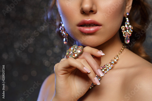 Woman and luxury jewelry in close-up. Glossy lips, elegant lady with manicured hand, crystal ring, necklace and earrings. Beauty model with healthy skin on dark bokeh background.