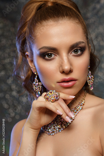 Woman and luxury jewelry in elegant portrait. Young lady holds her hand near her face, showing a large crystal ring, sparkling necklace and earrings. Beauty model on dark glitter bokeh background.