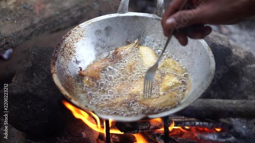 Close-up shot of a person's hand frying chicken pieces in hot bubbling oil using a traditional firewood stove.