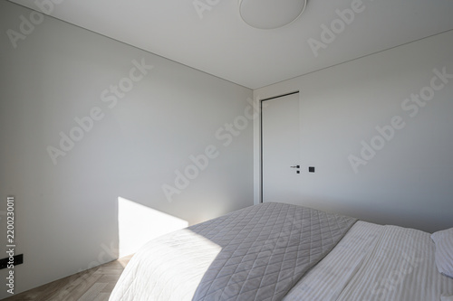 Minimalist bedroom features white quilted bed with striped sheets, flush-mount ceiling light, light wood floor, plain walls, subtle sunlight, clean geometry, soft shadows