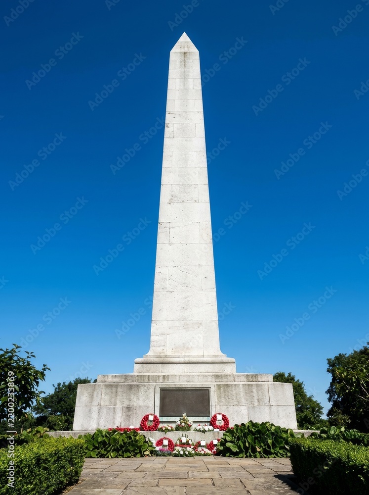 Fototapeta premium White stone obelisk war memorial against a clear blue sky with red poppy wreaths placed at the base